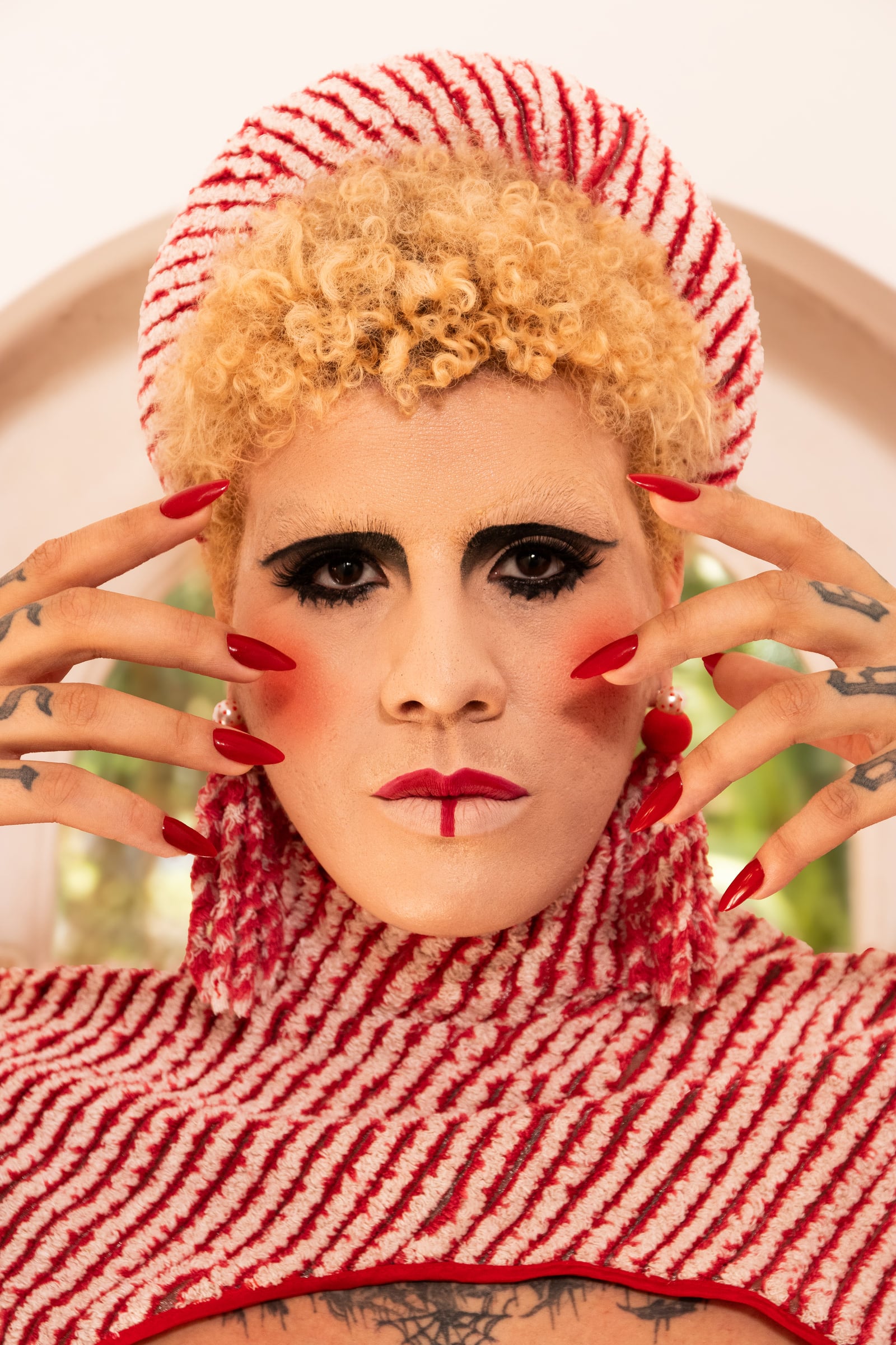 Frontal portrait with hands at the temples, red nails, black and red makeup and a striped fabric collar.